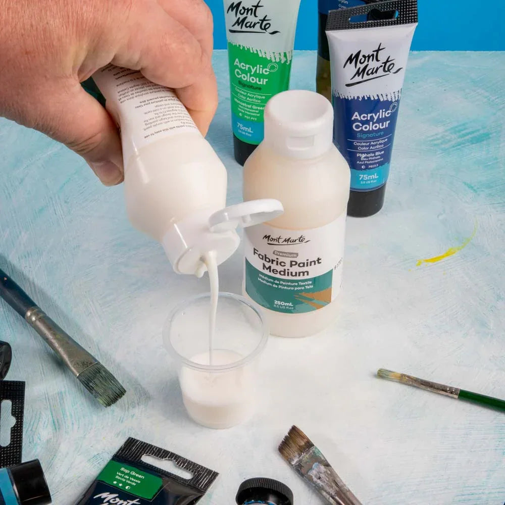Mont Marte Fabric Paint Medium being poured, surrounded by acrylic colours and paintbrushes on a blue surface.