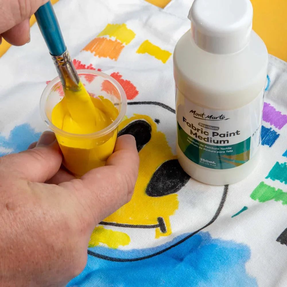 Mont Marte Fabric Paint Medium 250ml bottle beside hand painting colorful smiley face on fabric.
