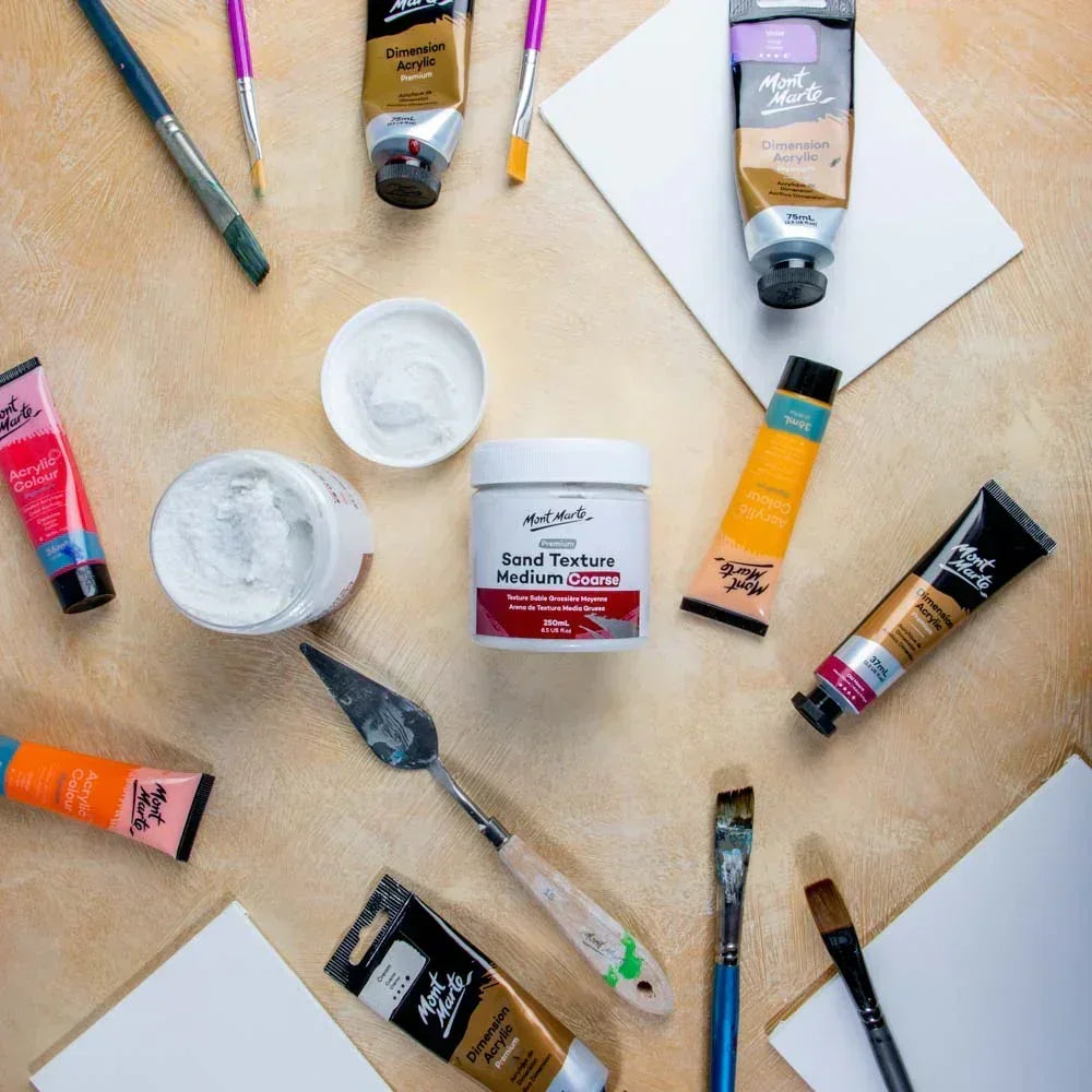 Mont Marte Sand Texture Medium Coarse 250ml with acrylic paints, palette knife, brushes, and canvases on a beige surface